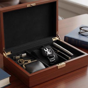 Puede incluir: Una caja de reloj de madera oscura, abierta para revelar un interior negro con un reloj negro con un dise&ntilde;o blanco, bol&iacute;grafos, un peque&ntilde;o monedero y otros accesorios. La caja est&aacute; sobre una superficie de madera.