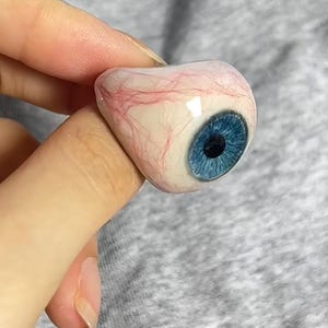 May include: A ring featuring a realistic, three-dimensional human eyeball design. The eyeball has a bright blue iris, a black pupil, and a white sclera detailed with fine, red blood vessels. The surface of the eye is smooth, glossy, and rounded.