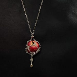 May include: Silver-toned necklace featuring a red apple pendant. The apple is encased in a decorative silver cage with sparkling accents. The necklace has a delicate chain and a small, ornate charm at the bottom.