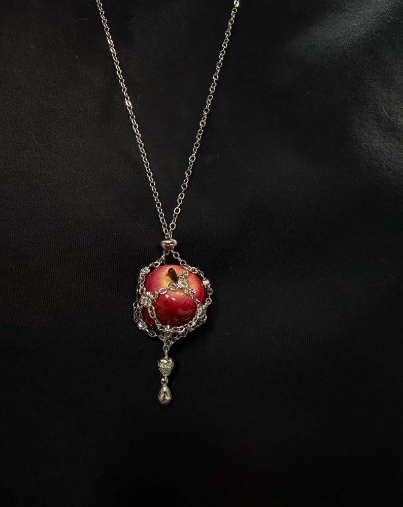 May include: Silver-toned necklace featuring a red apple pendant. The apple is encased in a decorative silver cage with sparkling accents. The necklace has a delicate chain and a small, ornate charm at the bottom.