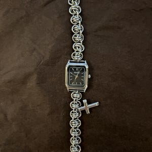 May include: Silver-toned watch with a black square face and a chain-link band. The watch has a small cross charm. The watch face has the word "NAIDU" printed on it.