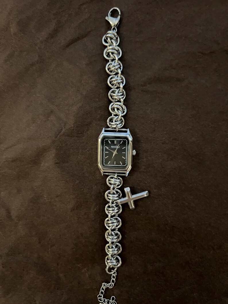May include: Silver-toned watch with a black square face and a chain-link band. The watch has a small cross charm. The watch face has the word "NAIDU" printed on it.