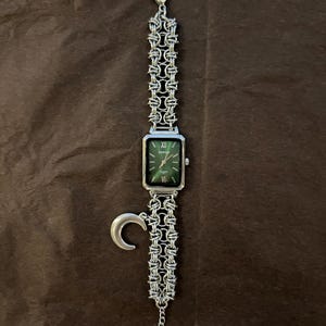 May include: Silver-toned watch with a rectangular green face and Roman numeral hour markers. The watch features a chain-link band and a crescent moon charm. The watch is displayed on a brown surface.