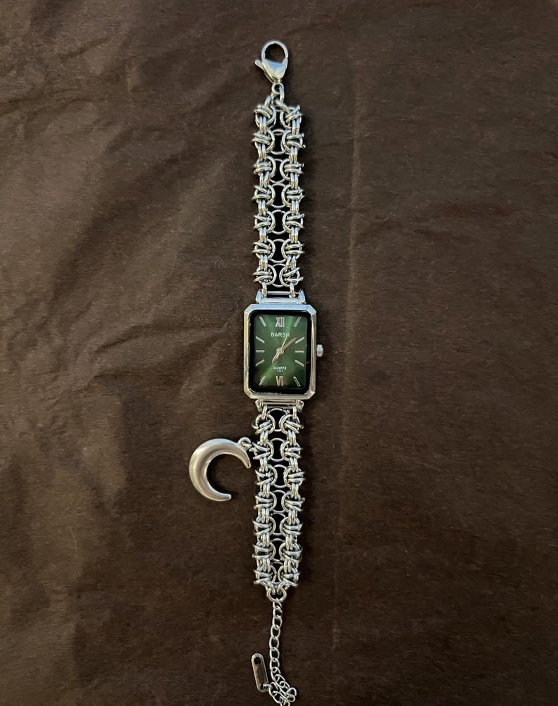 May include: Silver-toned watch with a rectangular green face and Roman numeral hour markers. The watch features a chain-link band and a crescent moon charm. The watch is displayed on a brown surface.