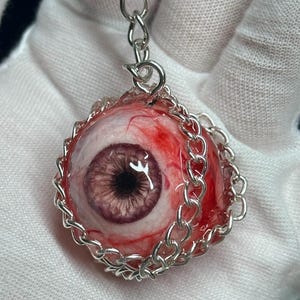 May include: A realistic eyeball pendant with a silver chain. The eyeball is detailed with red veins and a purple iris. The pendant is surrounded by a silver chain, creating a unique and macabre accessory.