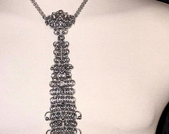 Könnte beinhalten: Eine silberfarbene Halskette aus Kettengeflecht mit einem krawattenartigen Anhänger. Die Halskette hat ein detailliertes, verziertes Design mit mehreren ineinandergreifenden Ringen. Der Anhänger hat einen dekorativen oberen Teil und einen langen, vertikalen Kettengeflechtteil.