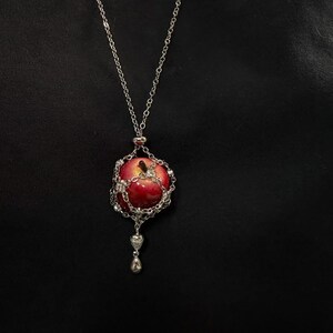 May include: A silver necklace featuring a red apple pendant. The apple is encased in a silver chain and adorned with clear crystals. A small silver heart and teardrop dangle below the apple, creating a unique and elegant accessory.