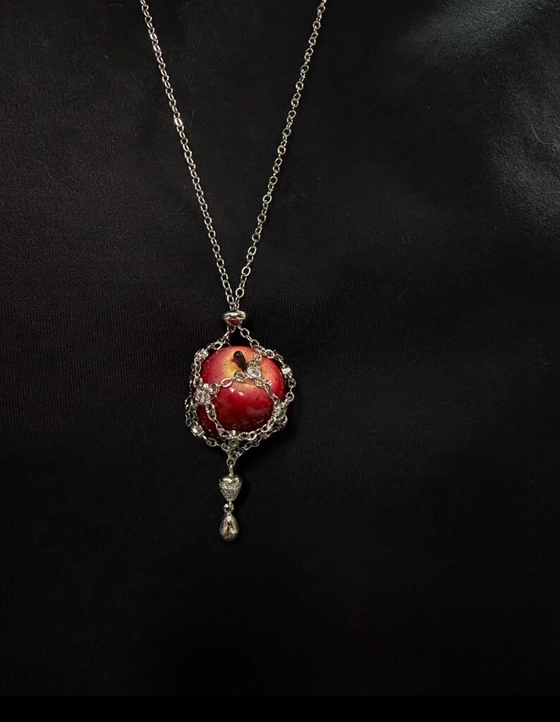 May include: A silver necklace featuring a red apple pendant. The apple is encased in a silver chain and adorned with clear crystals. A small silver heart and teardrop dangle below the apple, creating a unique and elegant accessory.