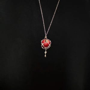 May include: A silver necklace with a red orb pendant. The pendant is a red sphere encased in a silver filigree cage, with a small silver charm dangling below. The chain is delicate and hangs against a black background.