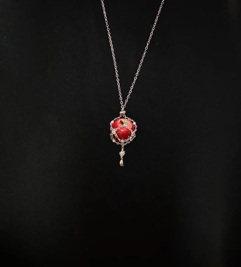 May include: A silver necklace with a red orb pendant. The pendant is a red sphere encased in a silver filigree cage, with a small silver charm dangling below. The chain is delicate and hangs against a black background.