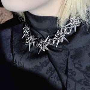 May include: A silver-colored spiked choker necklace. The necklace features a chain design with multiple sharp, conical spikes. The necklace is worn over a black garment with a subtle floral pattern.