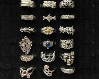 Puede incluir: Una colección de anillos de plata expuestos sobre una superficie negra. Los anillos presentan diversos diseños, incluyendo plumas, calaveras y patrones geométricos. Algunos anillos tienen piedras preciosas, otros detalles intrincados.