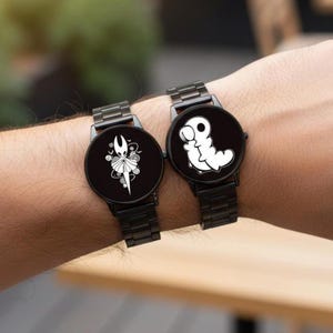 Puede incluir: Dos relojes de pulsera negros con correas de metal negro. Cada esfera del reloj presenta un dise&ntilde;o gr&aacute;fico blanco &uacute;nico. Un reloj muestra un personaje con corona, el otro una criatura blanca con forma de gusano. Los relojes se llevan en la mu&ntilde;eca.