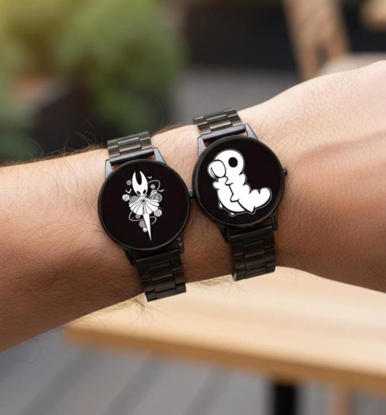 Puede incluir: Dos relojes de pulsera negros con correas de metal negro. Cada esfera del reloj presenta un dise&ntilde;o gr&aacute;fico blanco &uacute;nico. Un reloj muestra un personaje con corona, el otro una criatura blanca con forma de gusano. Los relojes se llevan en la mu&ntilde;eca.