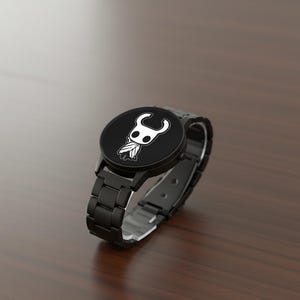 Puede incluir: Reloj de pulsera negro con una esfera redonda que presenta un gr&aacute;fico blanco de un personaje. El reloj tiene una correa de metal negro con cierre de hebilla. El reloj se muestra sobre una superficie de madera marr&oacute;n.