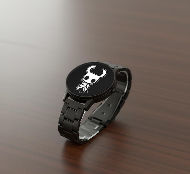 Puede incluir: Reloj de pulsera negro con una esfera redonda que presenta un gr&aacute;fico blanco de un personaje. El reloj tiene una correa de metal negro con cierre de hebilla. El reloj se muestra sobre una superficie de madera marr&oacute;n.