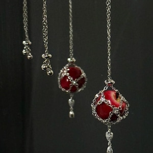 May include: Silver chain necklaces with red apple pendants. Each pendant features a red apple encased in a silver chain cage, with a dangling silver charm. The necklaces hang against a dark background.