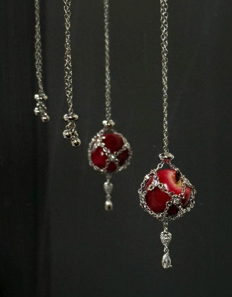 May include: Silver chain necklaces with red apple pendants. Each pendant features a red apple encased in a silver chain cage, with a dangling silver charm. The necklaces hang against a dark background.