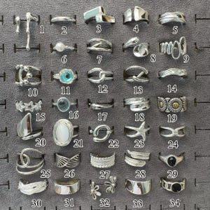 May include: A collection of 34 silver rings displayed on a gray surface. The rings feature various designs, including geometric shapes, pearls, gemstones, and floral motifs. Each ring is unique, showcasing diverse styles and textures.