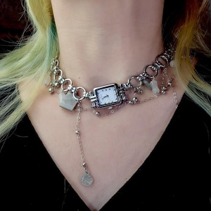 May include: A silver-toned watch necklace with a square watch face and a white dial. The necklace features a chain with dangling charms, including small beads and geometric shapes. The watch face displays the time. The necklace is worn around the neck.