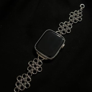 May include: Silver-toned Apple Watch with a chainmail-style band. The band features a floral pattern and a clasp closure. The watch face is black, and the band is made of a shiny metal. The watch is set against a black background.