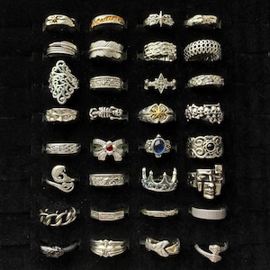 Może przedstawiać: Kolekcja srebrnych pierścionków o różnych wzorach, w tym filigranowych, piórkowych i geometrycznych. Niektóre pierścionki zdobią kamienie szlachetne, inne mają motywy czaszek, koron i łańcuchów. Pierścionki są prezentowane na czarnej powierzchni.