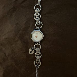 May include: A silver-toned wristwatch with an octagonal face and a white dial. The watch features a chain-link band and a small heart-shaped charm. The watch is displayed on a brown surface.