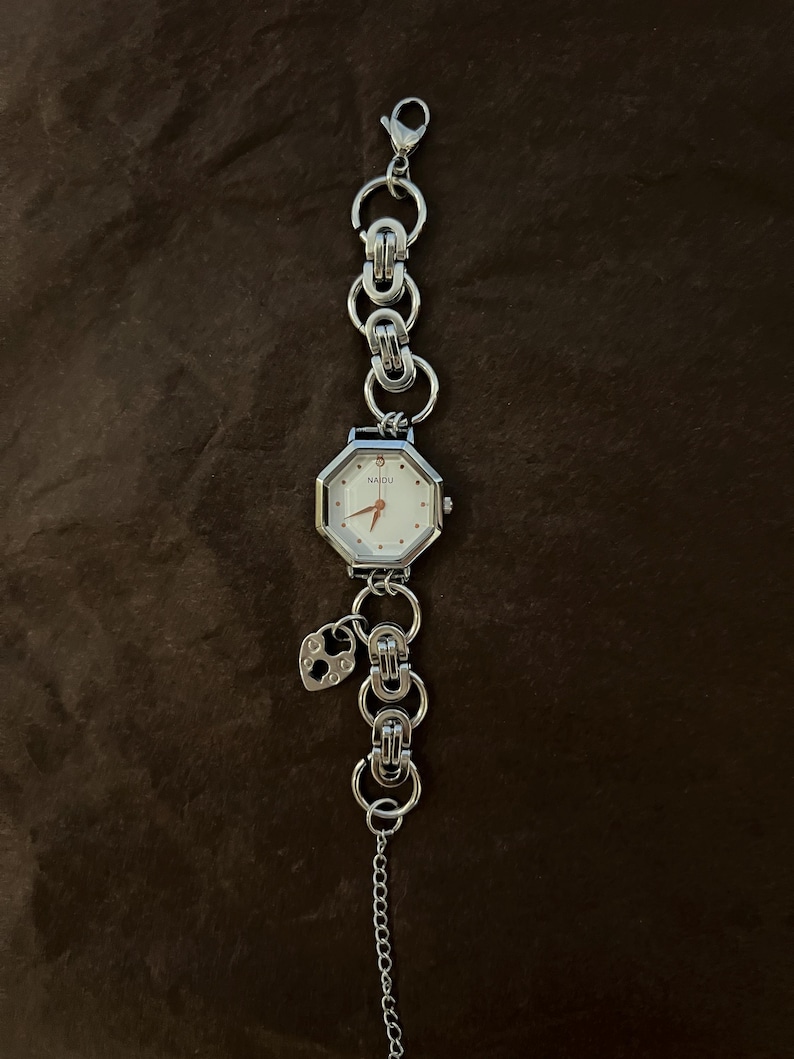May include: A silver-toned wristwatch with an octagonal face and a white dial. The watch features a chain-link band and a small heart-shaped charm. The watch is displayed on a brown surface.
