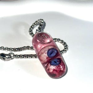 May include: A unique pendant necklace featuring a clear, capsule-shaped charm filled with pink resin and two blue eye-shaped inclusions. The pendant hangs from a silver-colored chain, creating a striking and unusual accessory.