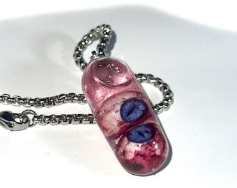 May include: A unique pendant necklace featuring a clear, capsule-shaped charm filled with pink resin and two blue eye-shaped inclusions. The pendant hangs from a silver-colored chain, creating a striking and unusual accessory.
