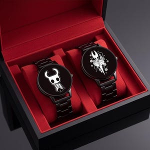 Puede incluir: Dos relojes de pulsera negros con correas de metal negro a juego se exhiben en una caja negra con interior de terciopelo rojo. Cada esfera del reloj presenta un diseño de personaje blanco sobre un fondo negro. Los relojes son un conjunto a juego.