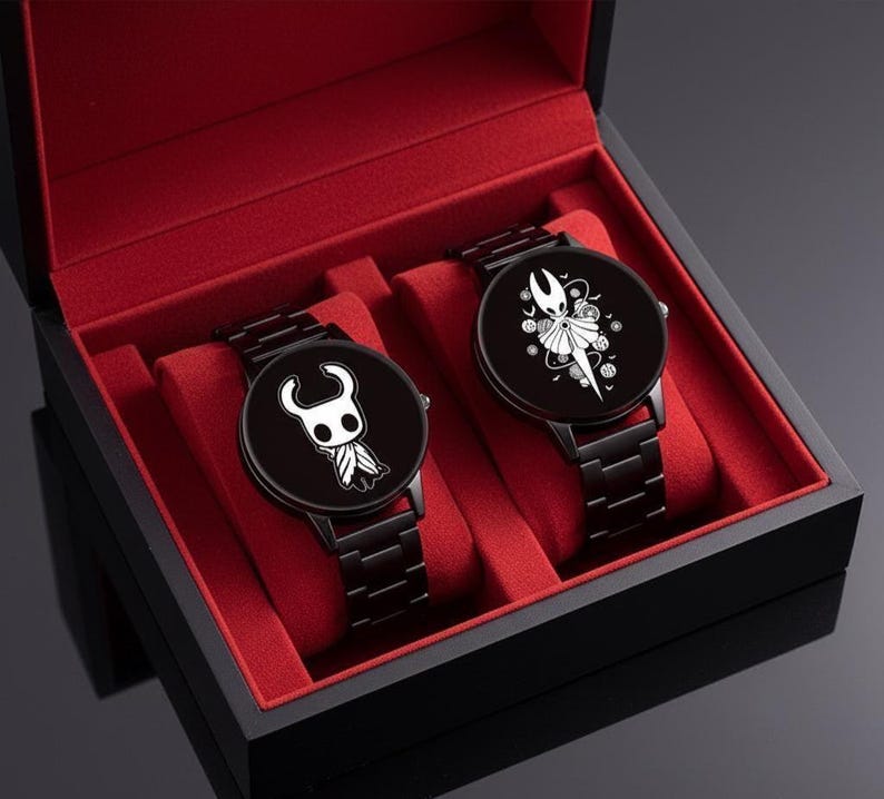Puede incluir: Dos relojes de pulsera negros con correas de metal negro a juego se exhiben en una caja negra con interior de terciopelo rojo. Cada esfera del reloj presenta un dise&ntilde;o de personaje blanco sobre un fondo negro. Los relojes son un conjunto a juego.