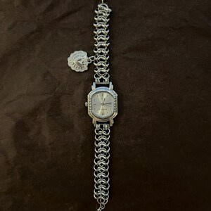 May include: Silver-toned wristwatch with a chain-link band and a decorative charm. The watch face is silver with a rectangular shape and a textured bezel. The band is made of interlocking metal links.