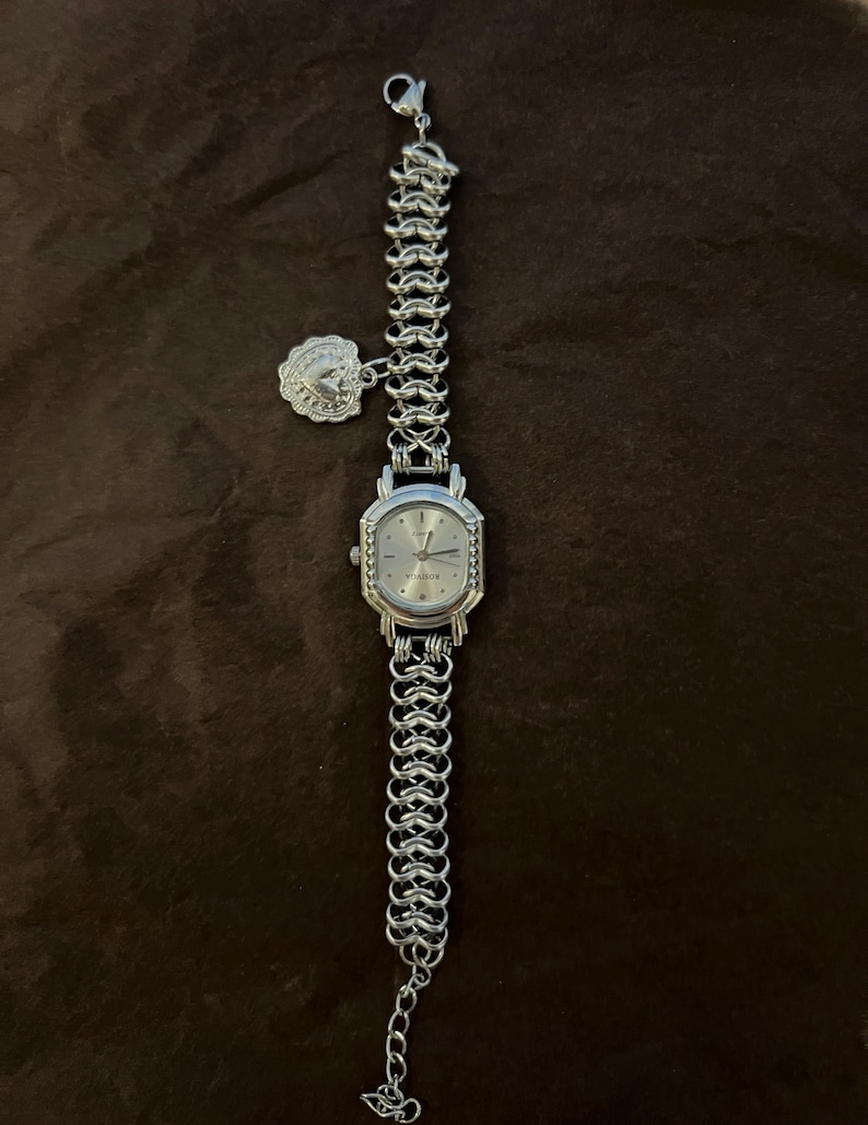 May include: Silver-toned wristwatch with a chain-link band and a decorative charm. The watch face is silver with a rectangular shape and a textured bezel. The band is made of interlocking metal links.