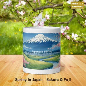May include: White ceramic mug featuring a scenic illustration of Mount Fuji with cherry blossoms and a river. The mug has the text "Japanese Spring" and "Spring in Japan - Sakura & Fuji". The background shows a green field and flowering tree branches.