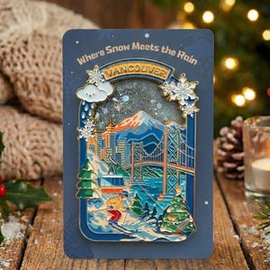 May include: A blue rectangular pin featuring a winter scene of Vancouver, with a bridge, mountains, and a skier. The text "Where Snow Meets the Rain" and "Vancouver" are displayed. Snowflakes and a cloud with rain complete the design.