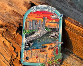 May include: A colorful enamel pin featuring a Vancouver cityscape with the word "Vancouver" at the top. The pin depicts a sunset, skyscrapers, a bridge, a ferry, and a volleyball game. A small airplane is also included.