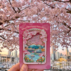 Pu&ograve; includere: Spilla smaltata rosa raffigurante un paesaggio urbano di Vancouver con fiori di ciliegio e la scritta "Where Blossoms Meet the Sea". La spilla include una catena montuosa, acqua ed edifici. Lo sfondo mostra un albero di fiori di ciliegio e uno skyline.