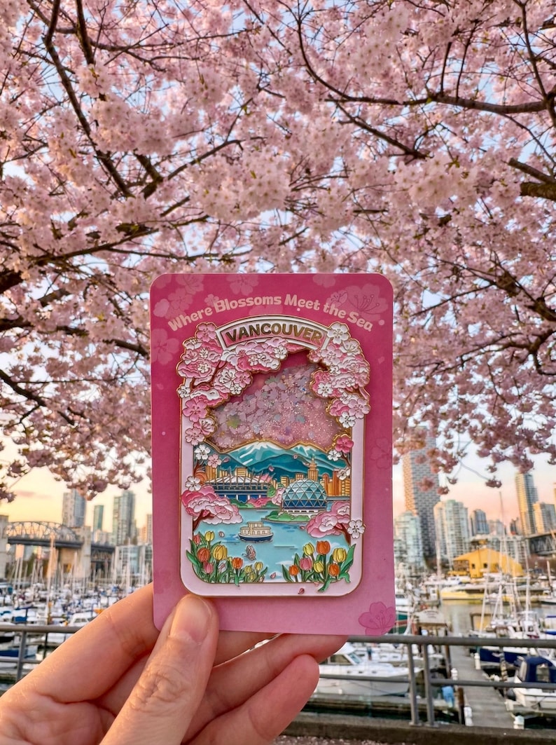 Pu&ograve; includere: Spilla smaltata rosa raffigurante un paesaggio urbano di Vancouver con fiori di ciliegio e la scritta "Where Blossoms Meet the Sea". La spilla include una catena montuosa, acqua ed edifici. Lo sfondo mostra un albero di fiori di ciliegio e uno skyline.