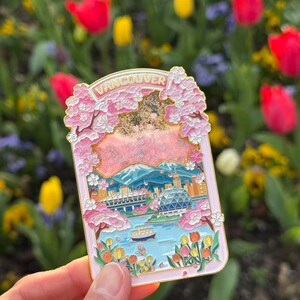 Pu&ograve; includere: Una spilla in smalto rosa raffigurante un paesaggio di Vancouver con fiori di ciliegio e lo skyline della citt&agrave;. La spilla ha la scritta "Vancouver" in alto e un bordo dorato. Lo sfondo &egrave; un giardino sfocato con tulipani rossi e gialli.