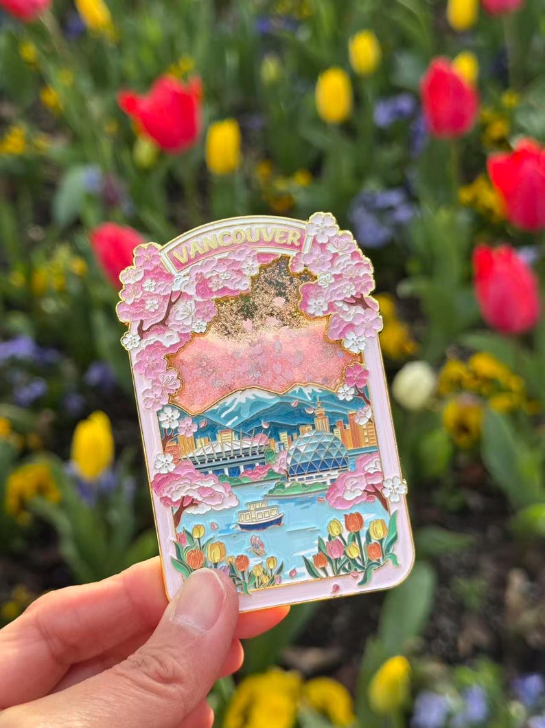Pu&ograve; includere: Una spilla in smalto rosa raffigurante un paesaggio di Vancouver con fiori di ciliegio e lo skyline della citt&agrave;. La spilla ha la scritta "Vancouver" in alto e un bordo dorato. Lo sfondo &egrave; un giardino sfocato con tulipani rossi e gialli.