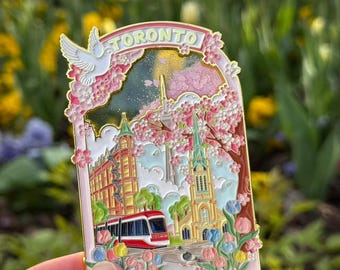 May include: A pink Toronto souvenir pin depicting a cityscape with a red and white tram, buildings, and a church. The pin has a floral border with the word "TORONTO" and a white dove.