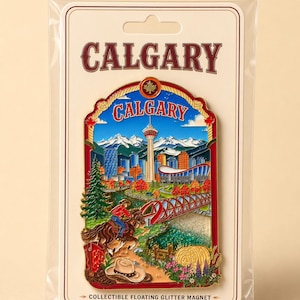 May include: A collectible floating glitter magnet featuring a colorful enamel design of the Calgary skyline, including the Calgary Tower, mountains, a red bridge, a person on horseback, a cowboy hat, and a red boot, all on a branded display card.