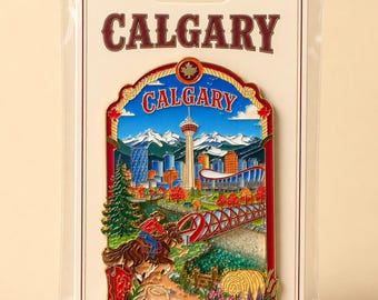May include: A collectible floating glitter magnet featuring a colourful enamel design of the Calgary skyline, including the Calgary Tower, mountains, a red bridge, a person on horseback, a cowboy hat, and a red boot, all on a branded display card.