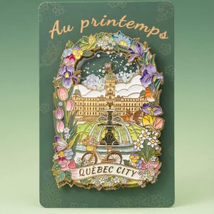 May include: A decorative pin featuring a colorful illustration of Quebec City, with a floral border and the words "Au printemps" and "Quebec City". The pin includes a bicycle, fountain, and building.