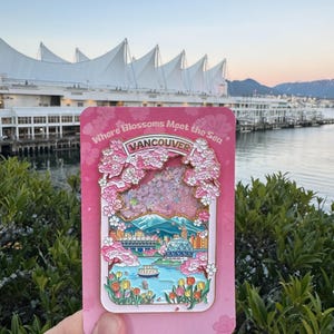 Pu&ograve; includere: Una spilla in smalto rosa raffigurante un paesaggio urbano di Vancouver con fiori di ciliegio e la scritta "Where Blossoms Meet the Sea" e "Vancouver". La spilla &egrave; tenuta davanti a una scena sul lungomare con una struttura bianca a forma di vela sullo sfondo.