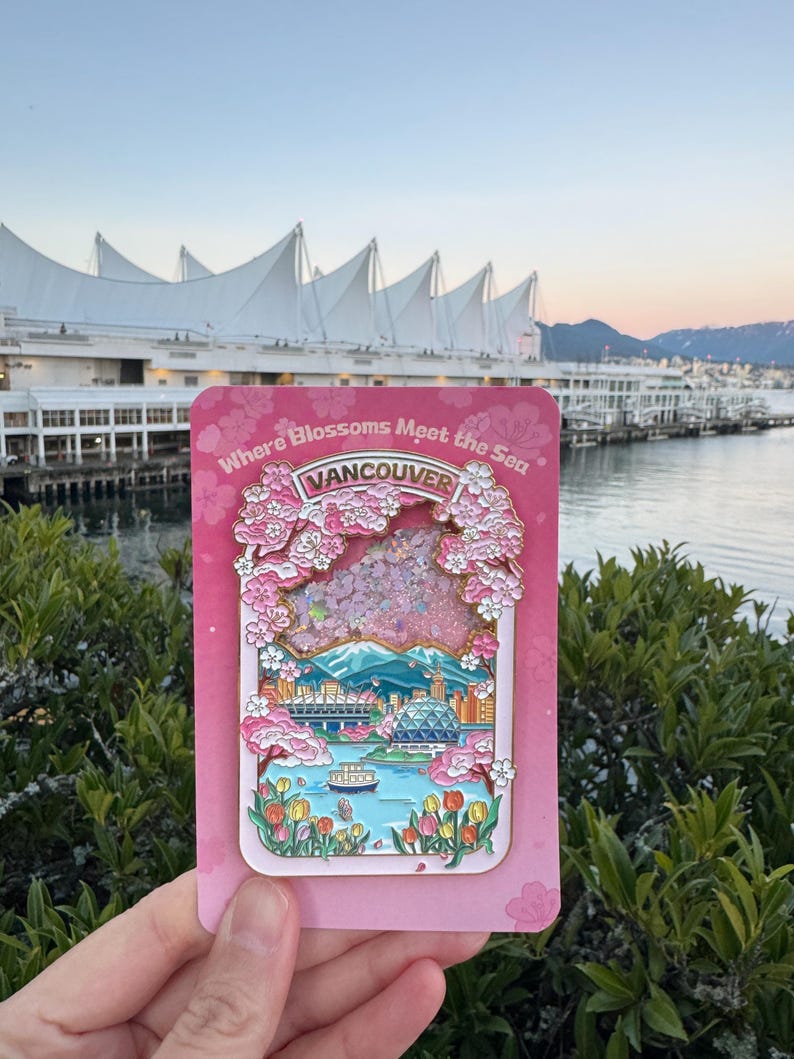 Pu&ograve; includere: Una spilla in smalto rosa raffigurante un paesaggio urbano di Vancouver con fiori di ciliegio e la scritta "Where Blossoms Meet the Sea" e "Vancouver". La spilla &egrave; tenuta davanti a una scena sul lungomare con una struttura bianca a forma di vela sullo sfondo.
