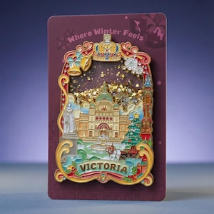 May include: A decorative enamel pin featuring a detailed scene of Victoria with a central building, a horse-drawn carriage, and a Christmas tree. The pin is set against a dark red backing card with the text Where Winter Feels at the top.