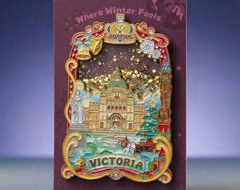 May include: A decorative enamel pin featuring a detailed scene of Victoria with a central building, a horse-drawn carriage, and a Christmas tree. The pin is set against a dark red backing card with the text Where Winter Feels at the top.