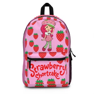 May include: Pink backpack featuring the Strawberry Shortcake character and red strawberries. The backpack has a black zipper and straps, with the words "Strawberry Shortcake" in red script. The design includes a cartoon illustration of Strawberry Shortcake.